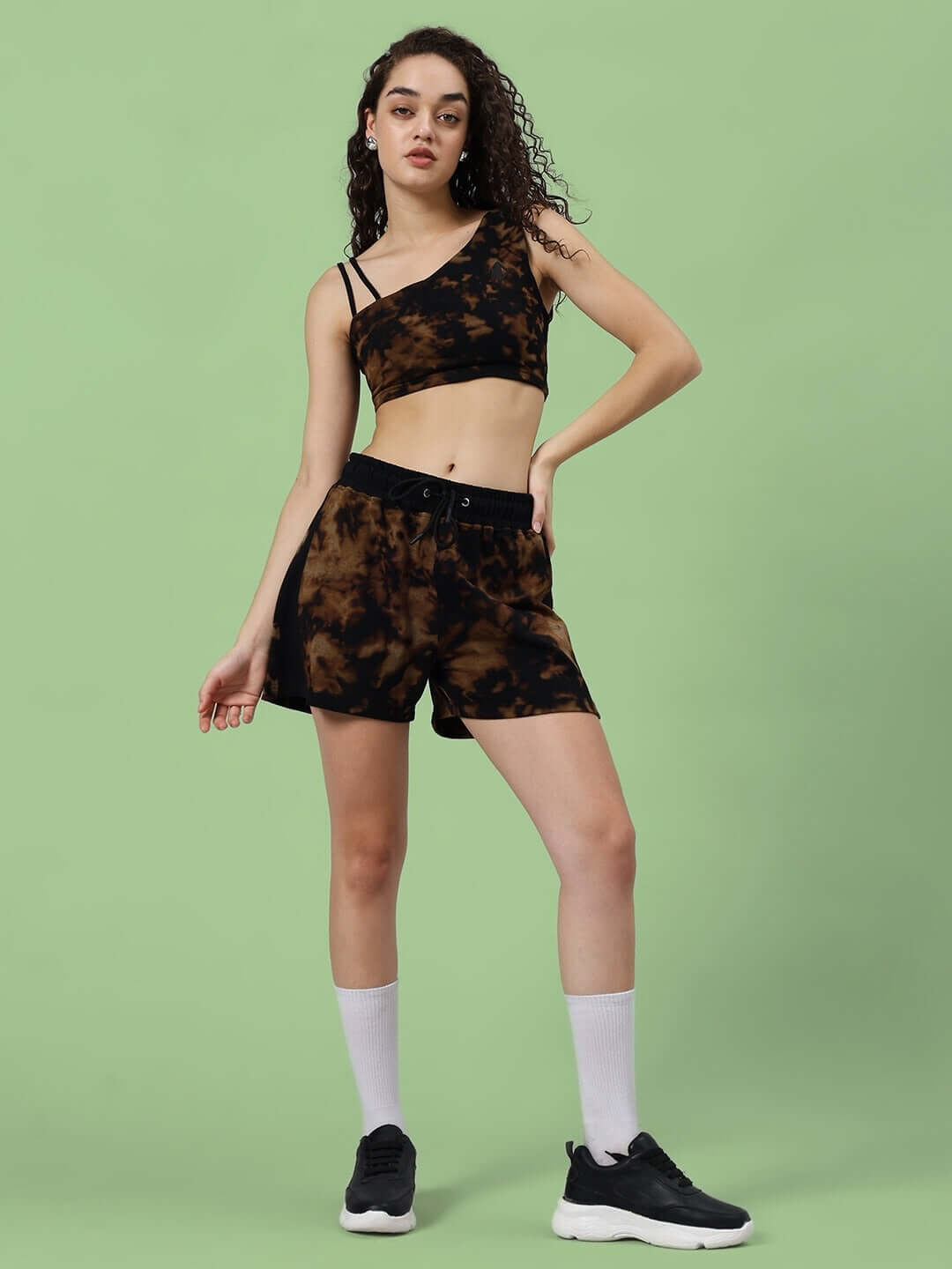 Woman wearing black tie-dye co-ord set with shorts and crop top, styled with white socks and black sneakers, green background.