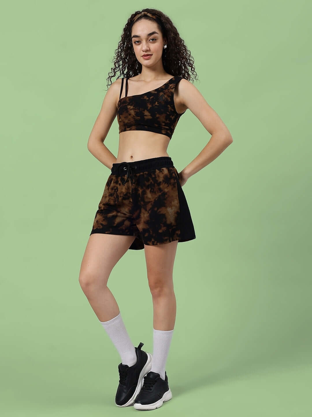 Woman wearing black tie-dye co-ord set featuring crop top and shorts, showcasing a blend of style and comfort, standing on green background.