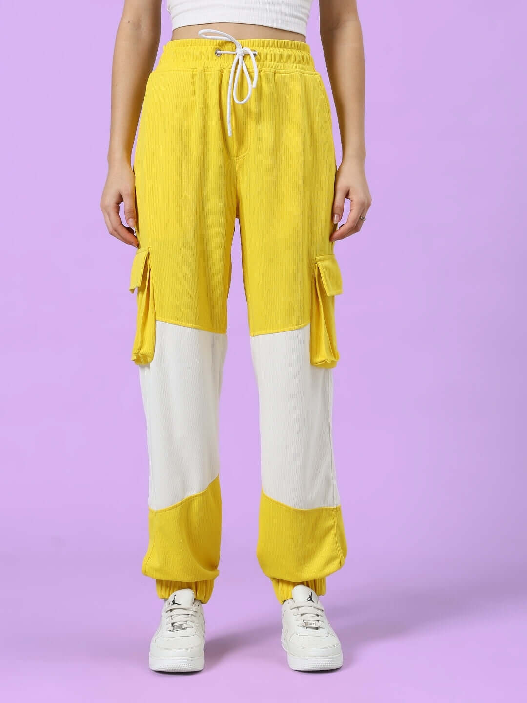 Woman wearing yellow and white relaxed fit joggers, featuring cargo pockets, against a purple background.