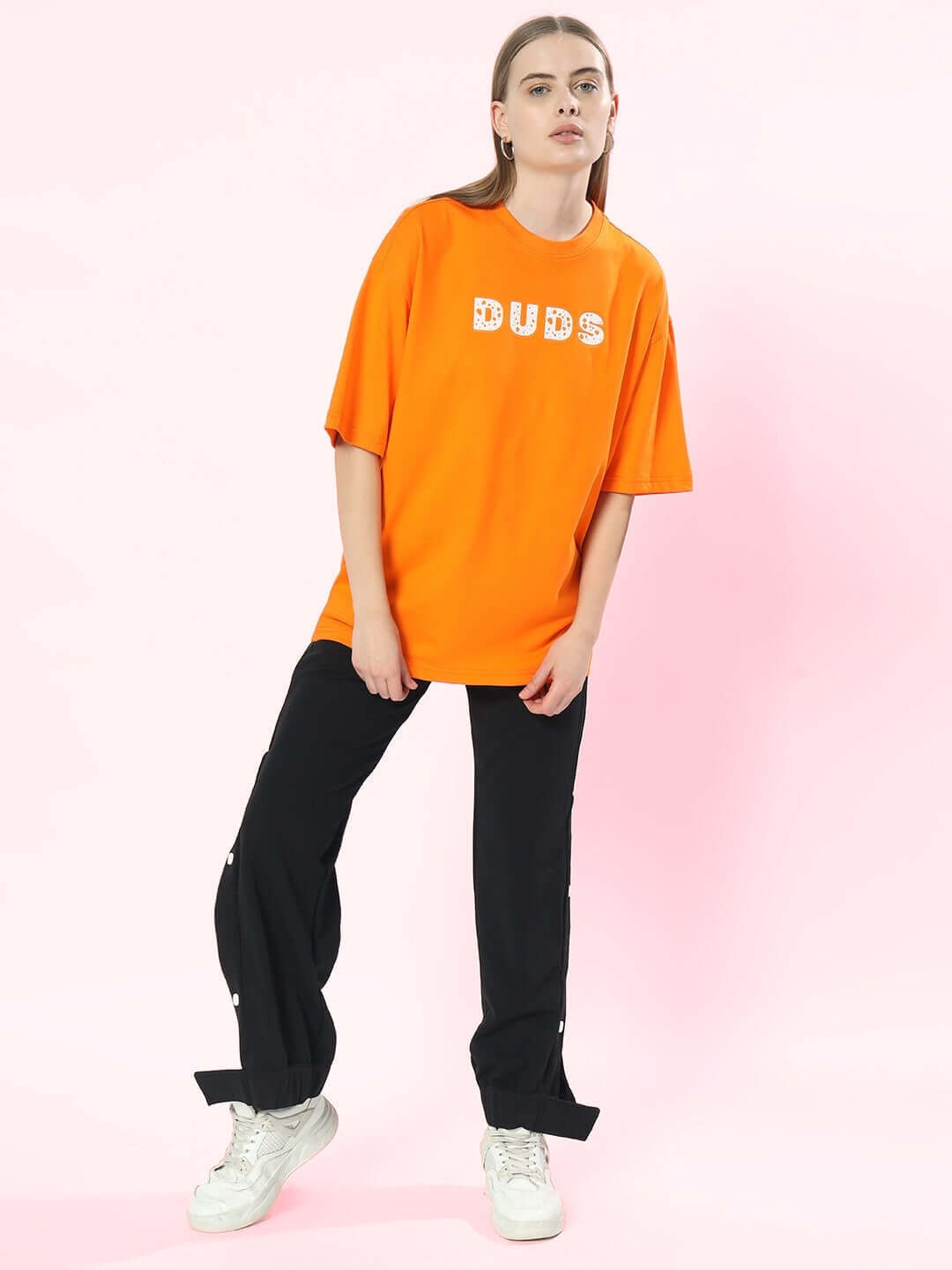 Model wearing DUDS orange oversized t-shirt with text, paired with black pants and white sneakers, on a pink backdrop.