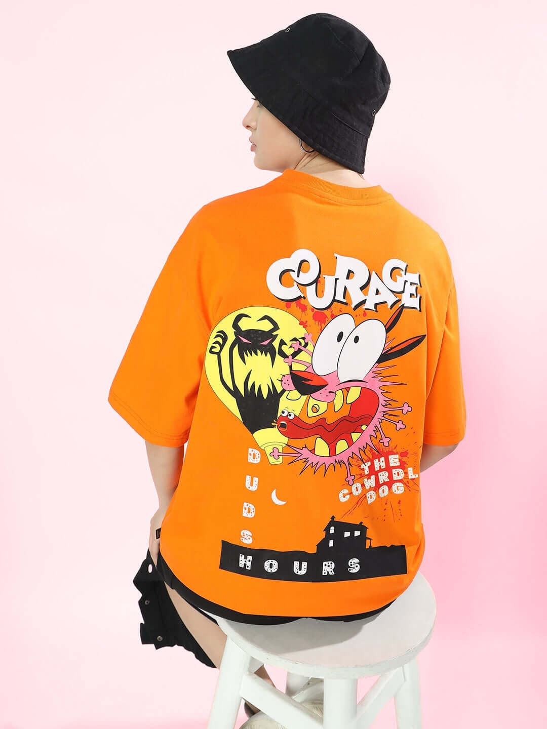 Woman wearing an oversized orange t-shirt with colorful graphics and 'Courage' text, paired with a black bucket hat, sitting on a stool.