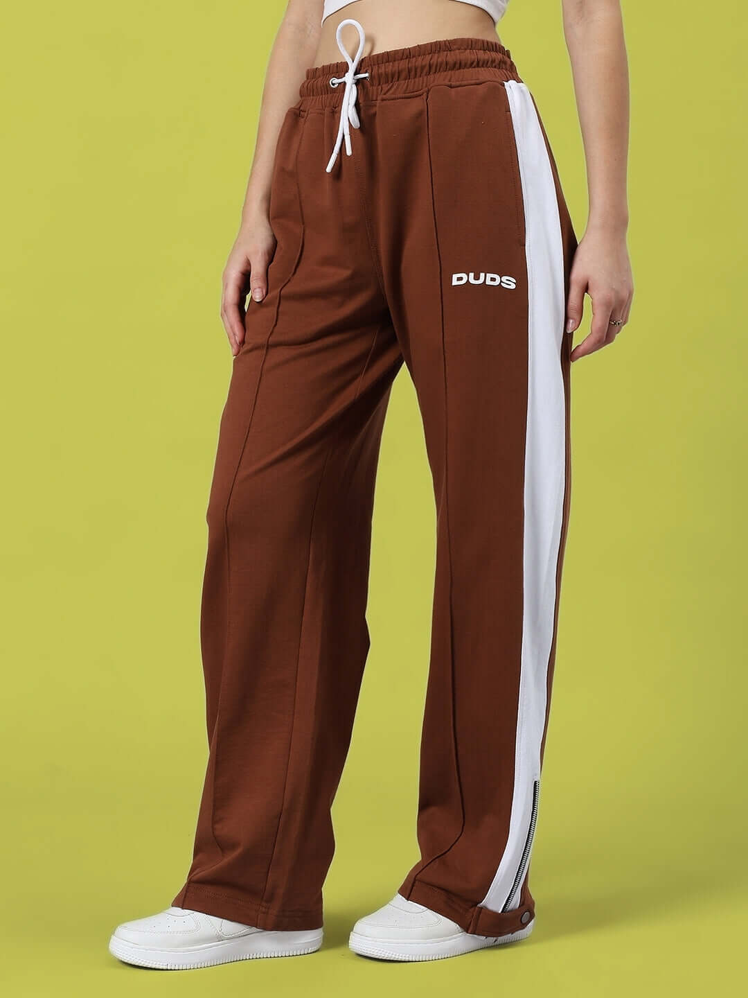 Women's brown joggers with slit zippers and white stripe from DUDS, shown on model standing against a green background.