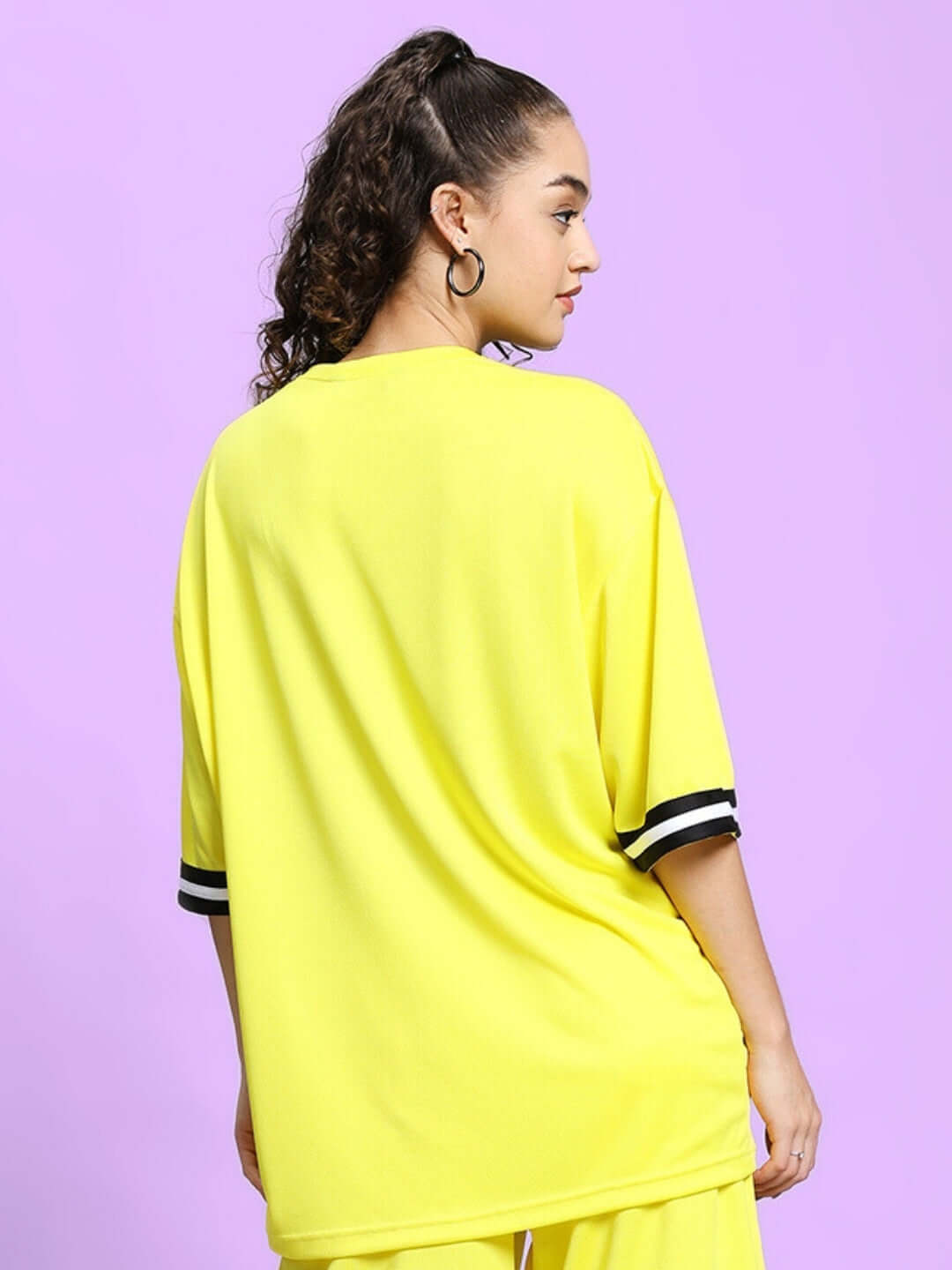 Woman wearing yellow oversized T-shirt with black and white striped sleeves, viewed from the back against a purple background.