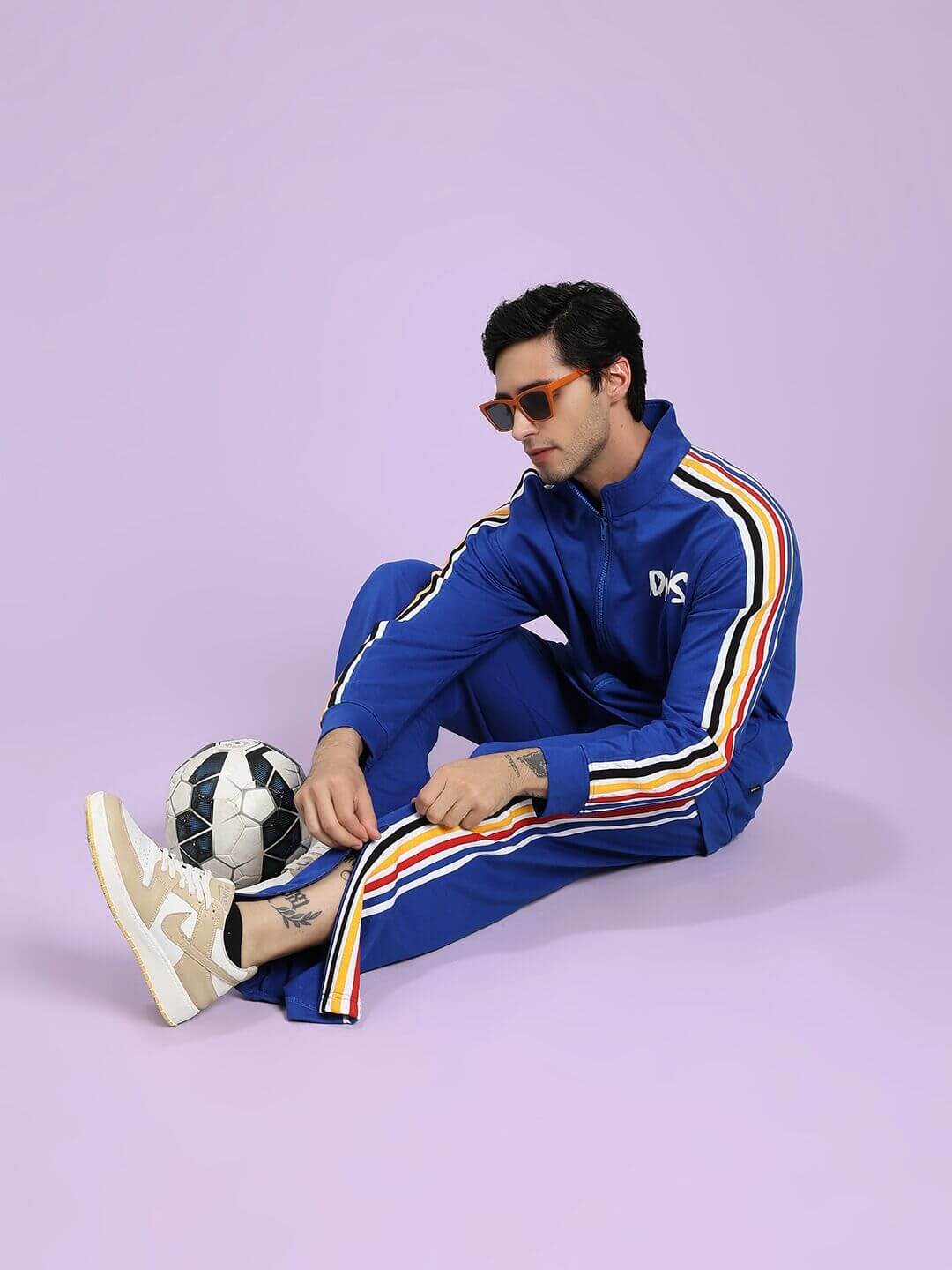 Person wearing a blue tracksuit with colorful stripes, sitting on the floor with a soccer ball against a light purple background.