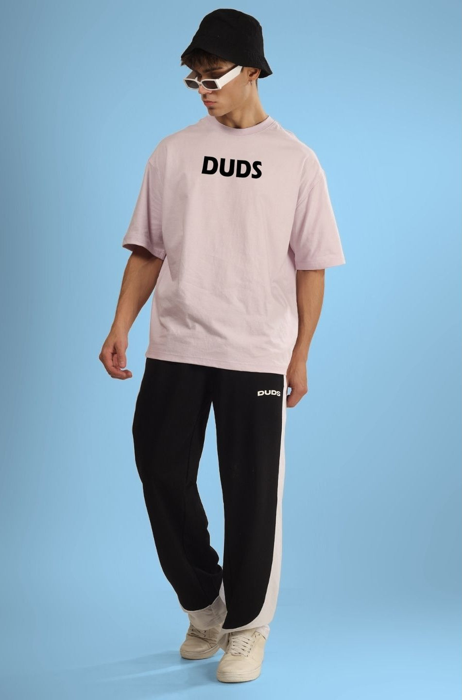 Man wearing STAY WILD OVERSIZED T-SHIRT GREY with black pants, bucket hat, and sunglasses, in stylish casual outfit on blue background.