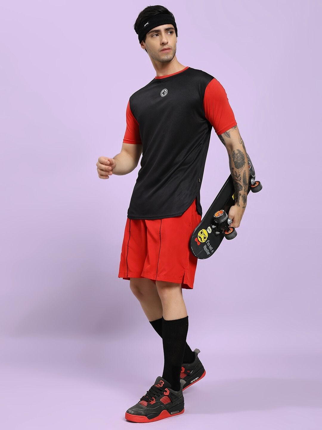 Man wearing a black and red athletic outfit holding a skateboard against a light purple background