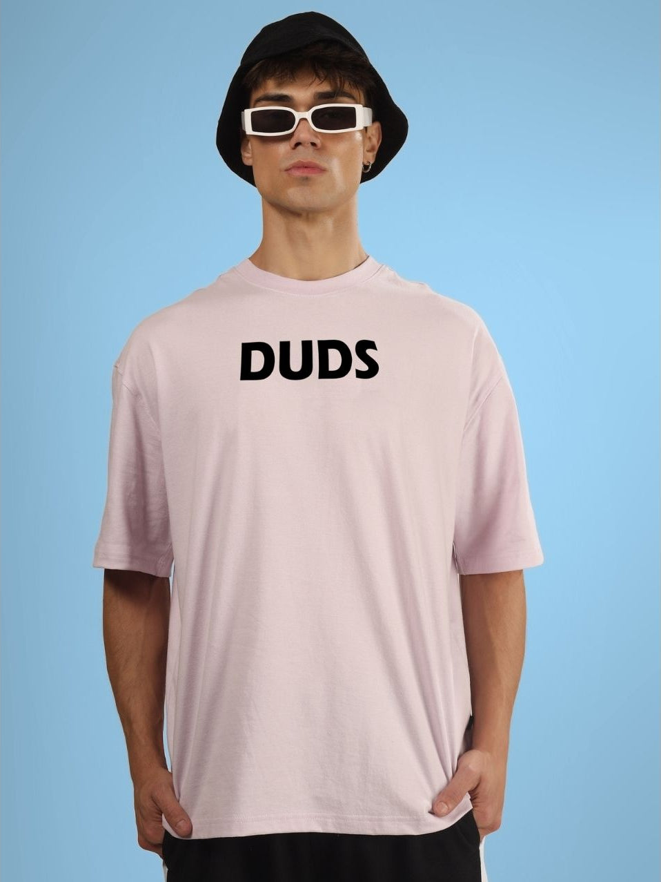 Model wearing a pink oversized t-shirt with 'DUDS' text, sunglasses, and a black hat against a blue background.