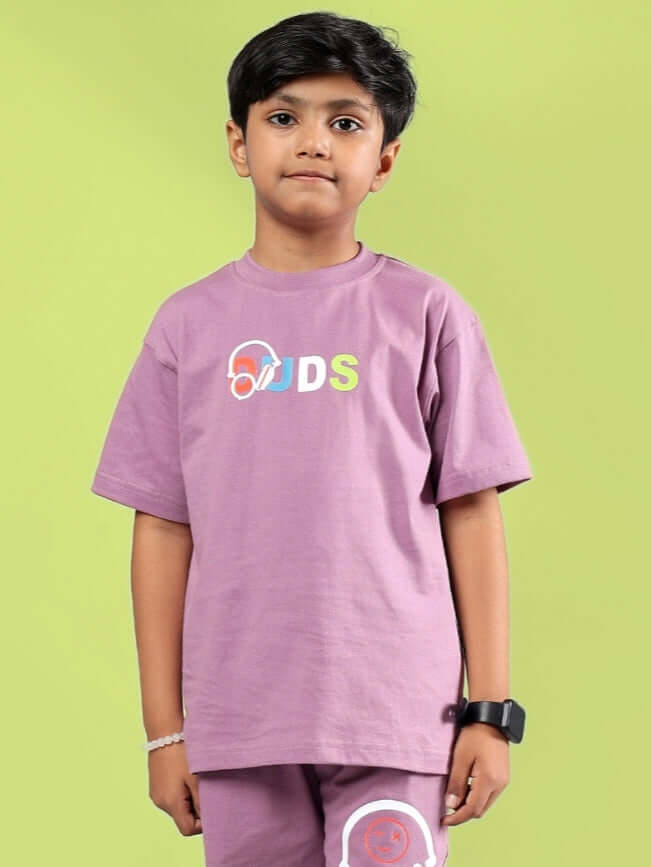 Child wearing Stoic T-shirt in Pulm Purple by DUDS with colorful logo on chest, set against a green background.
