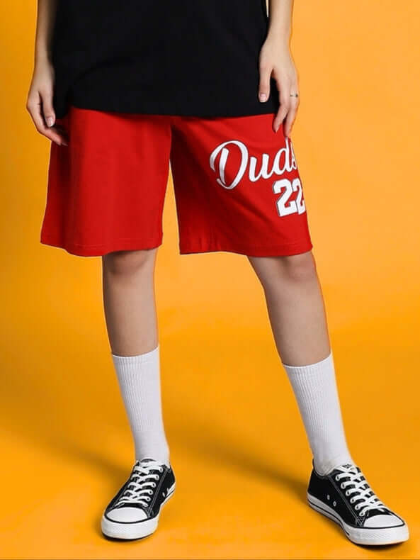 Women's red Columbia 22 regular fit shorts, featuring 'Duds 22' text, styled with white socks and black sneakers against an orange backdrop.