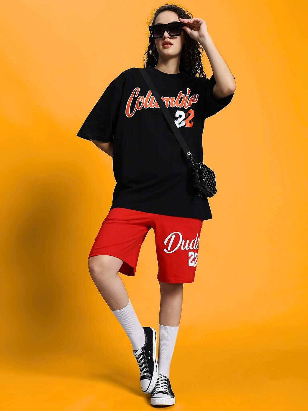 Woman wearing red Columbia 22 regular fit shorts with a black top, accessorized with sunglasses and a sling bag on an orange background.