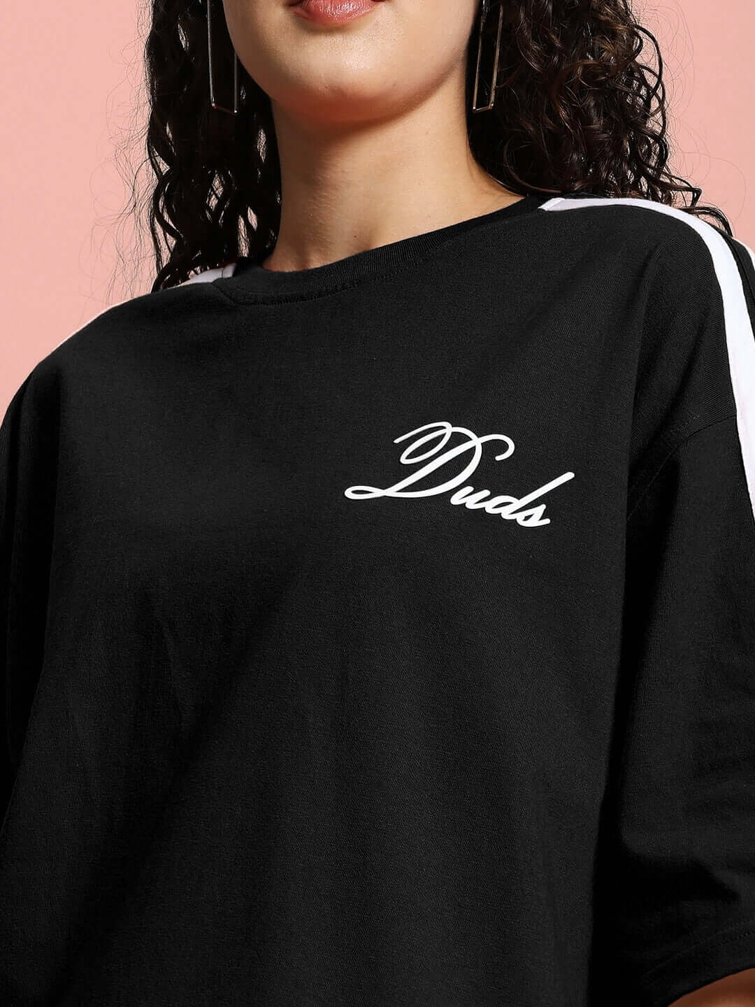Woman wearing black oversized t-shirt with 'Duds' logo, made from premium cotton blend fabric, featured against a pink background.
