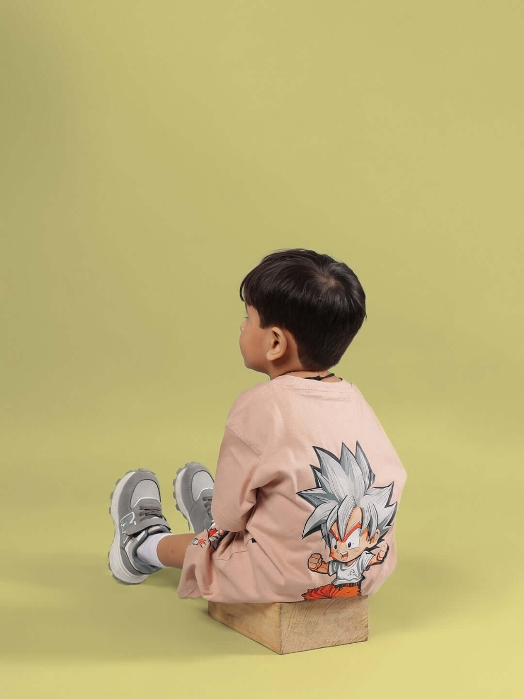 Child wearing a beige Gohan t-shirt, sitting on a box against a green background, displaying a cartoon character design on the back.