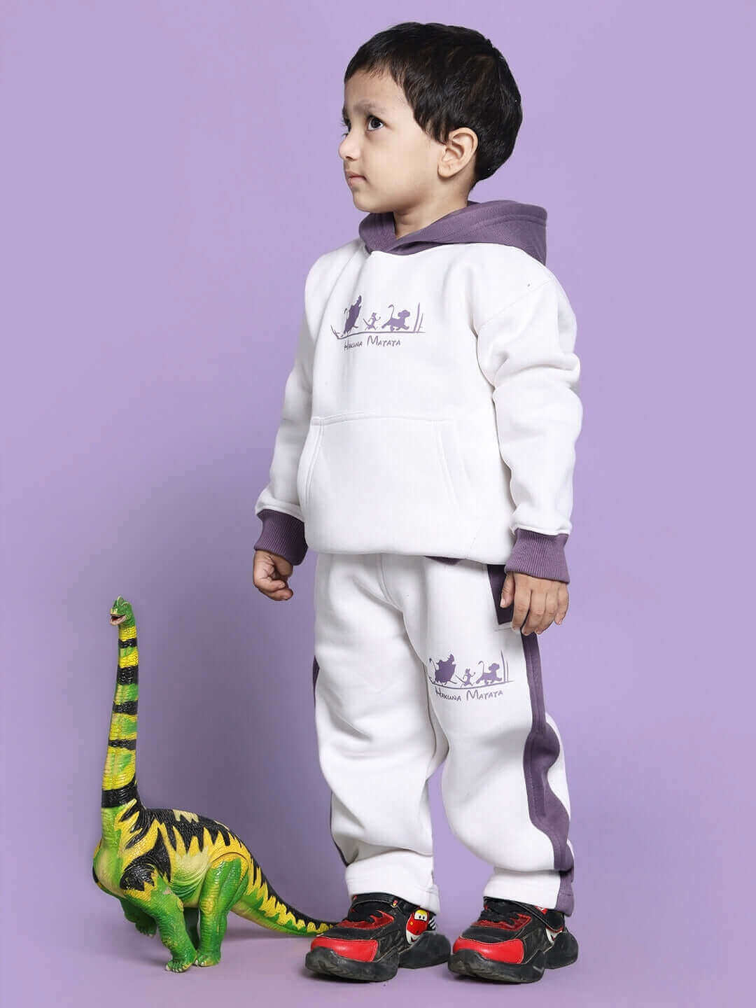 Child wearing Hakuna Matata white-violet fleece pants and hoodie set, standing beside a toy dinosaur, purple background.
