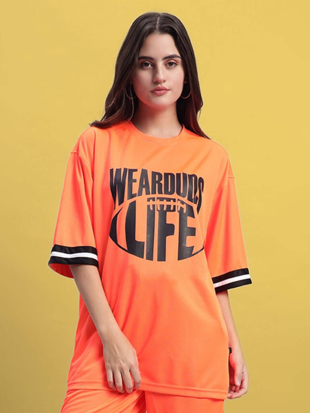 Woman wearing orange oversized T-shirt with 'WearDuds Life' text, featuring black stripes on sleeves, against yellow background.