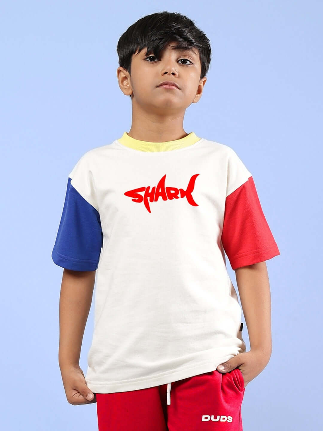 Boy wearing an oversized off-white T-shirt with shark print, featuring blue and red sleeves, designed for boys and girls by DUDS.