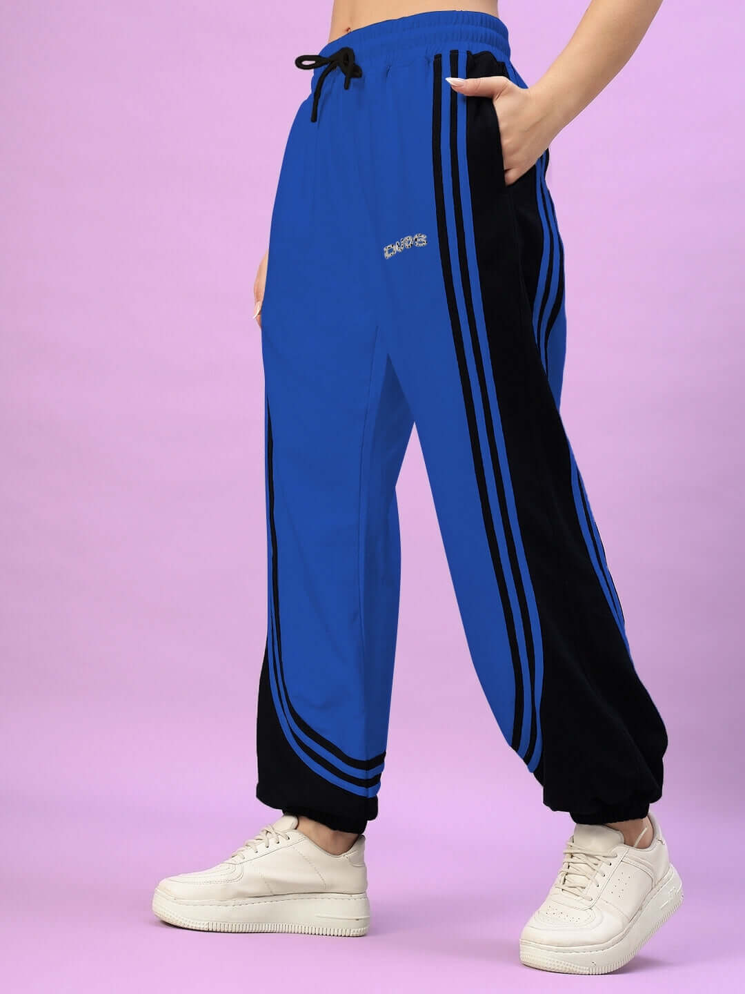 Blue and black women's joggers with contrast stripes, styled with white sneakers on a pink background. Trendy casual athletic wear.