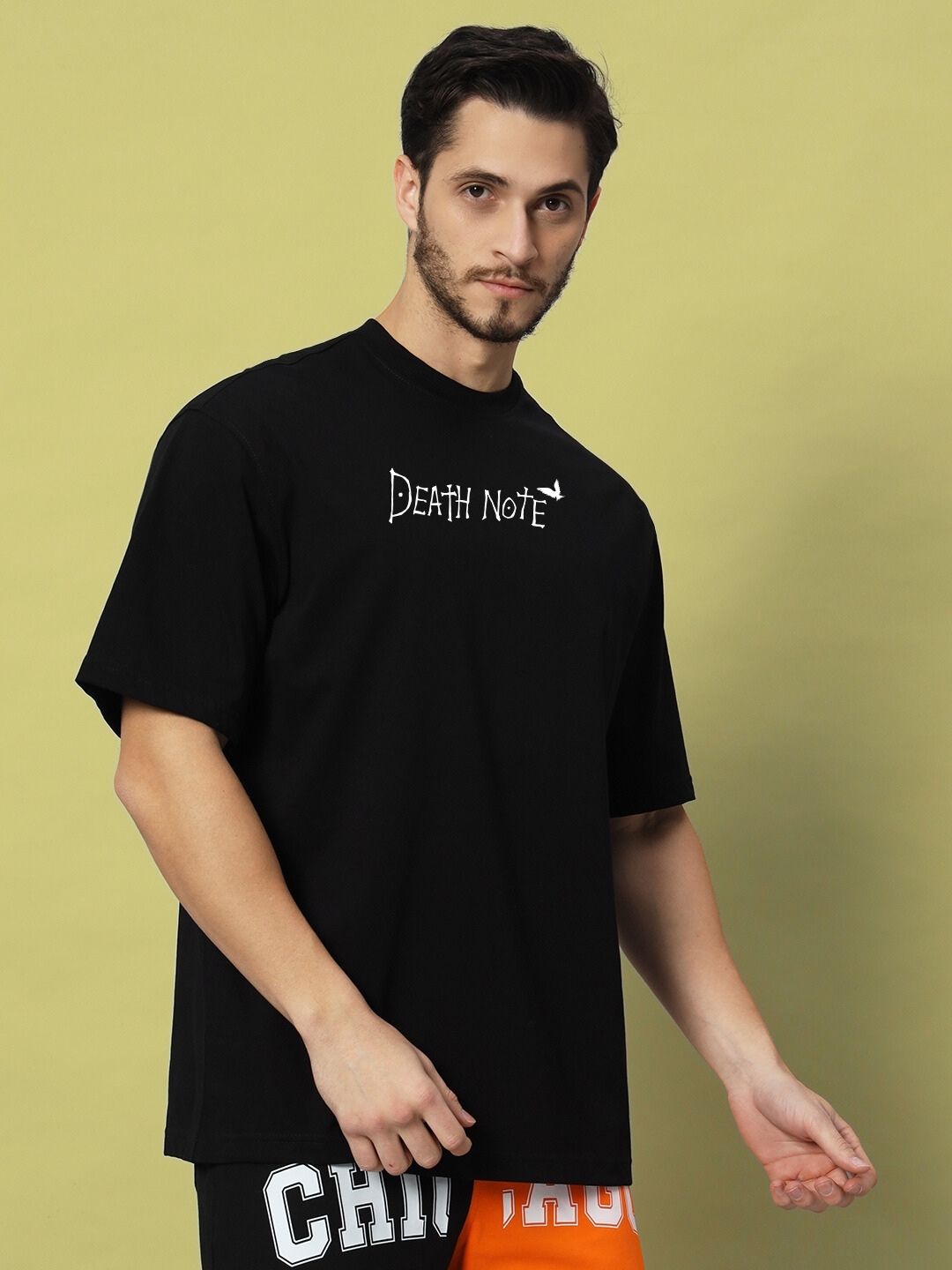 Man wearing FOLLOW LIFE OVERSIZED T-SHIRT BLACK, styled with casual printed shorts, standing against a light green background