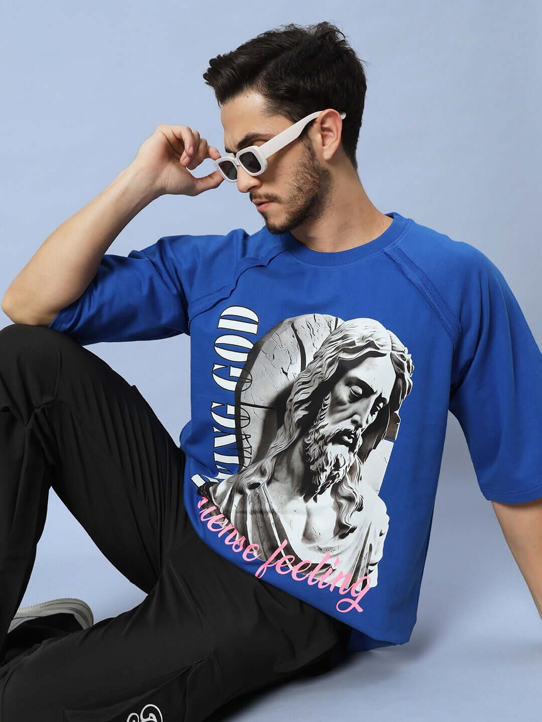 Man wearing REGLAN DYING GOD OVER-SIZED T-SHIRT ROYAL BLUE with graphic design, sitting in a relaxed pose against a light blue background.