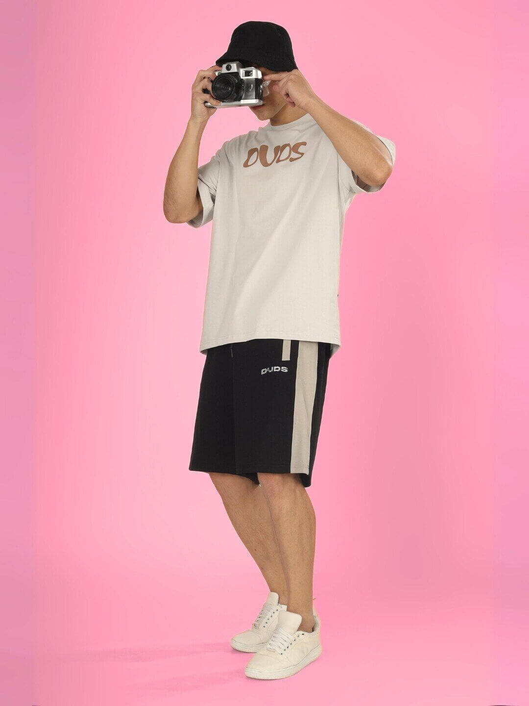 Model wearing HIGH AS FUCK OVER-SIZED T-SHIRT BUTTER CREAM with a camera, capturing chic streetwear style against a pink background.