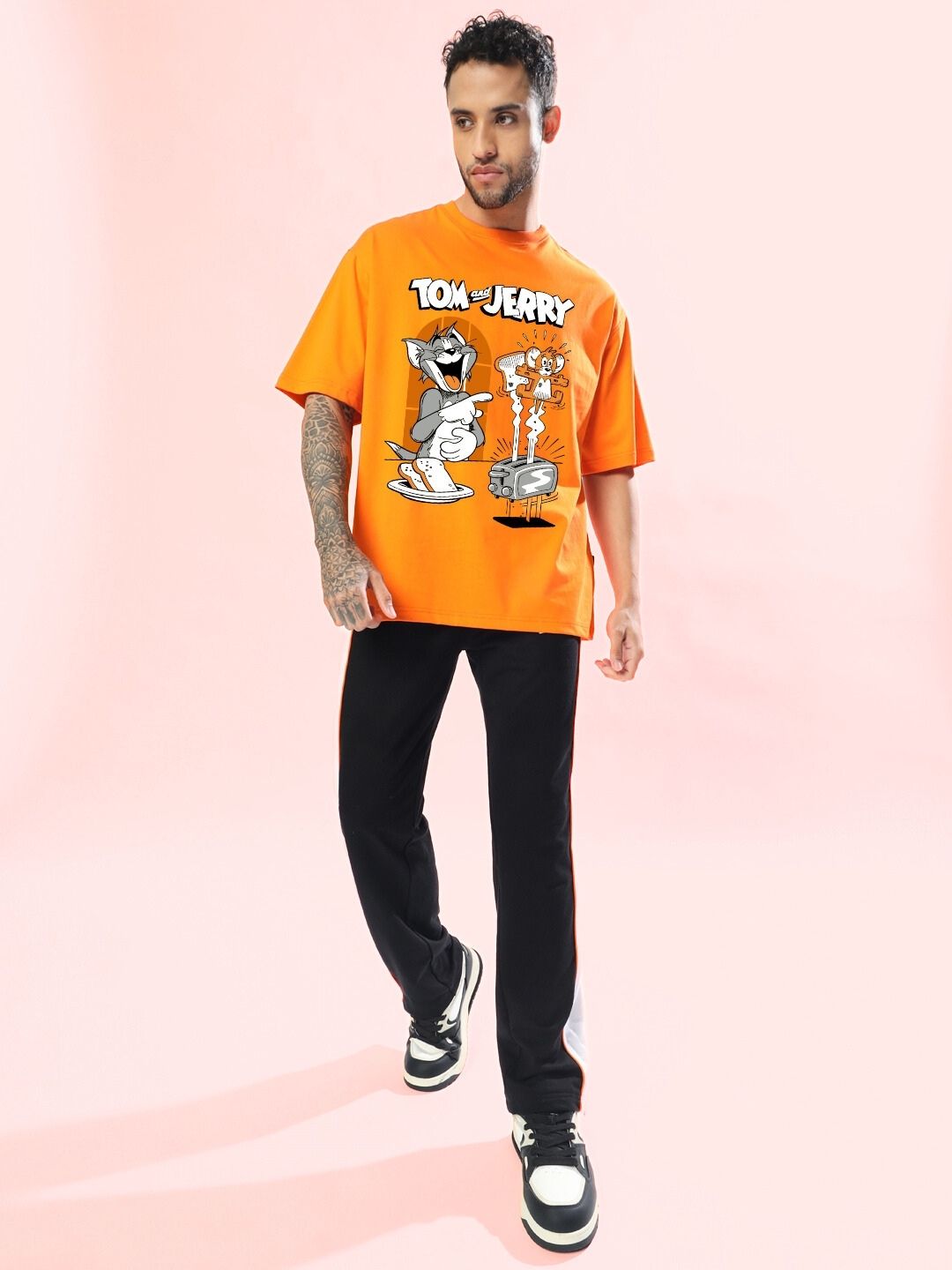 Man wearing TOM AND JERRY TOAST OVERSIZED T-SHIRT ORANGE with black pants against a pink background.