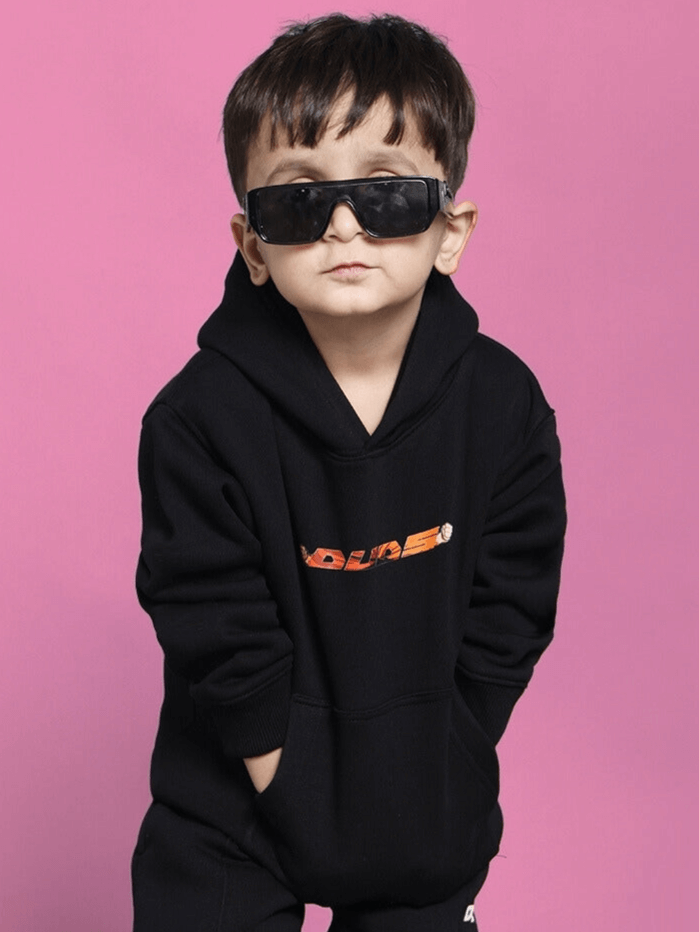 Child wearing black GOHAN hoodie with orange graphic, hands in pockets, and sunglasses, against a pink background.
