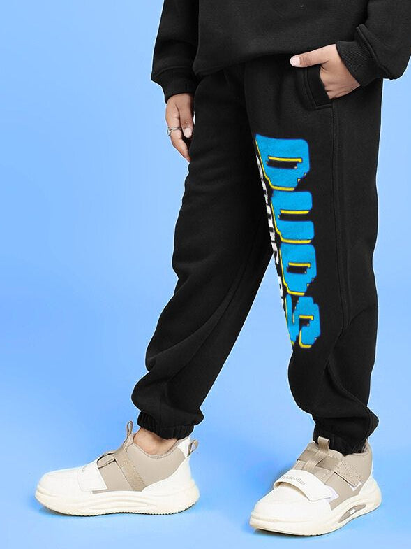 Child wearing GAME ON PRINT FLEECE JOGGER FOR BOYS AND GIRLS BLACK, featuring blue graphic design, stands with hands in pockets on blue background.