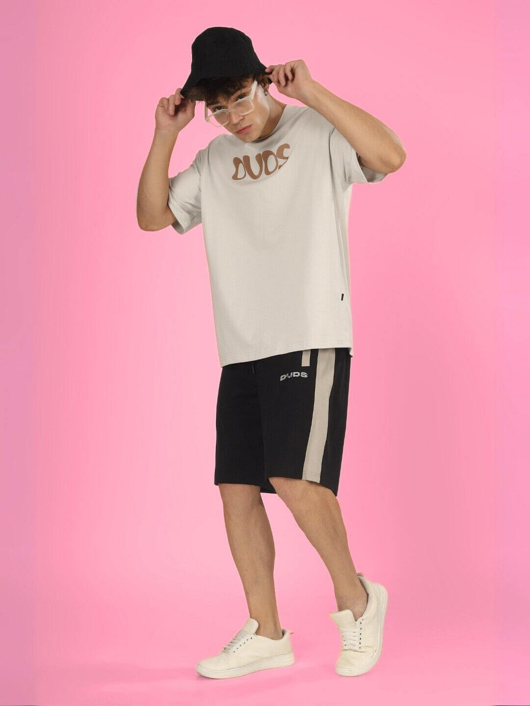 Model wearing HIGH AS FUCK OVER-SIZED T-SHIRT BUTTER CREAM with black cap and shorts, standing against pink background, embodying streetwear style.