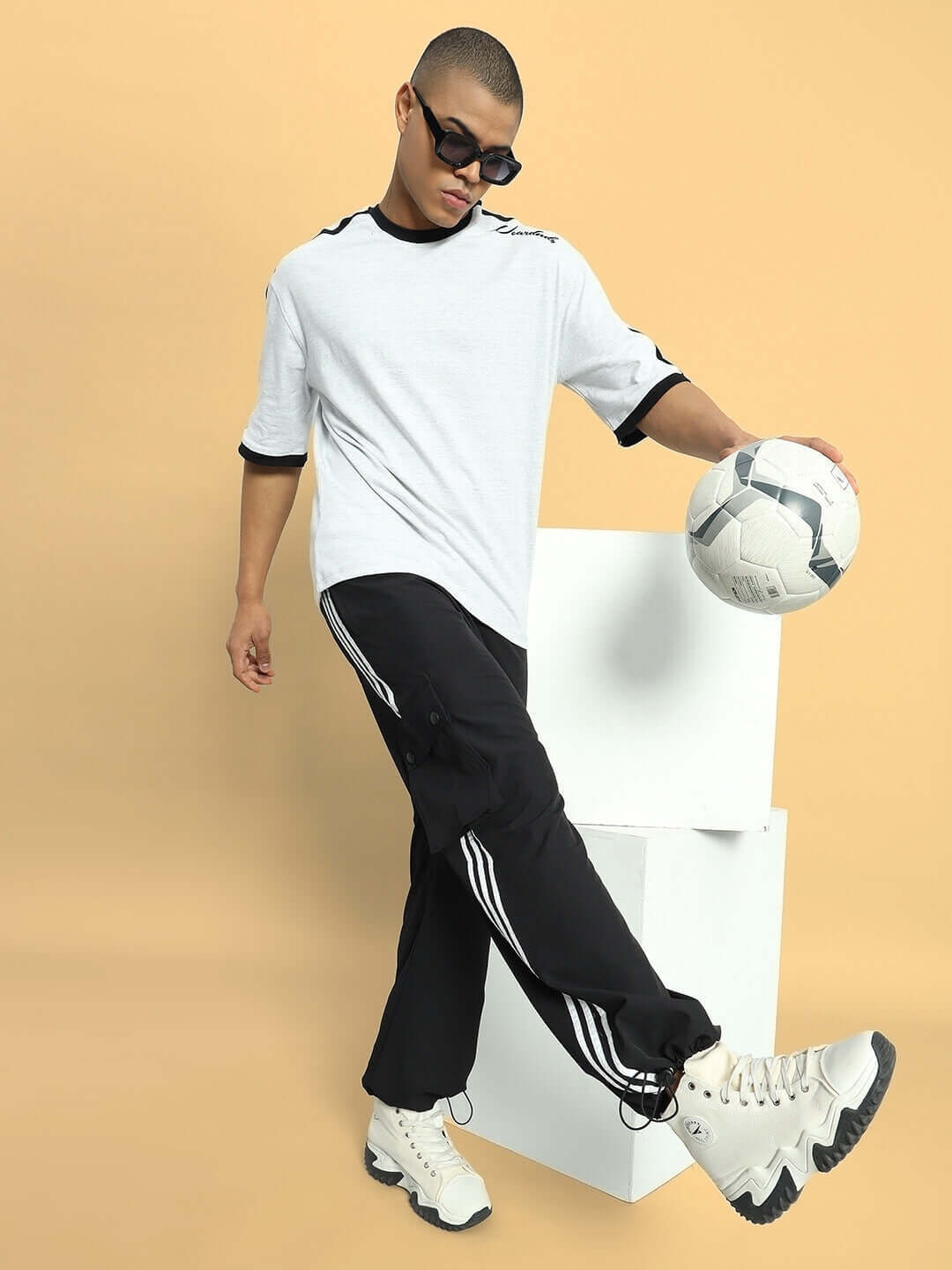 Model wearing NOVA Relaxed Fit Cargo Pants in black, paired with a white t-shirt and sneakers, holding a soccer ball for a sporty look.