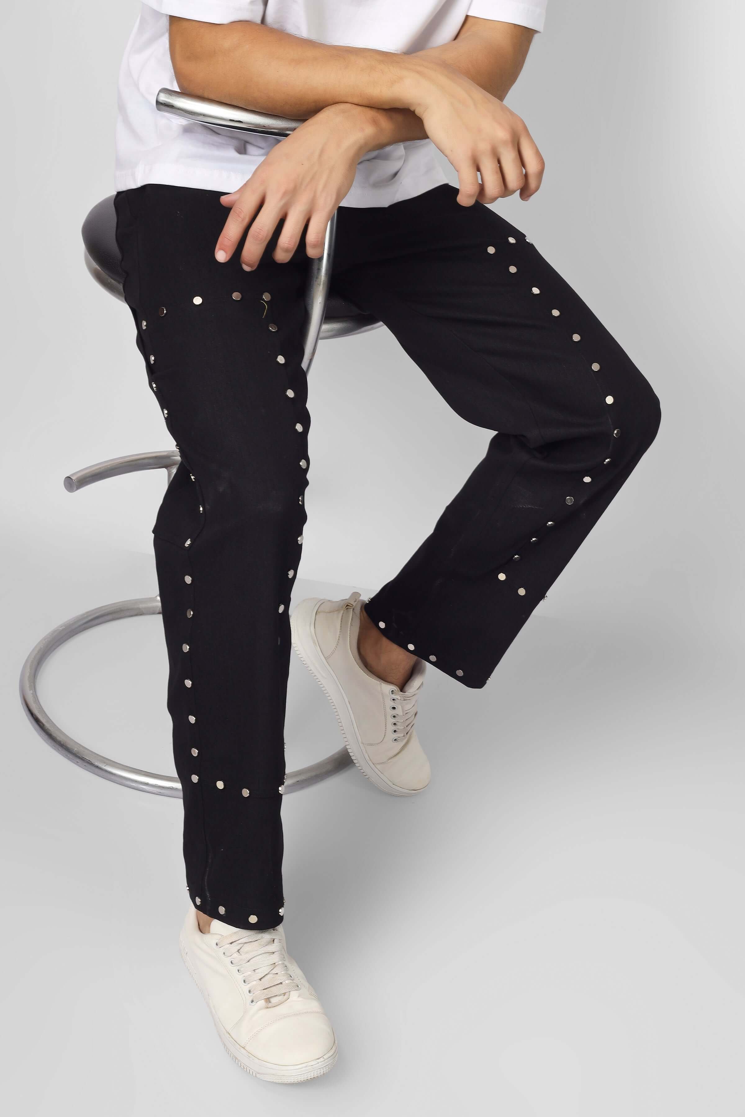 Model wearing black 7-pocket pants with metal stud buttons, sitting on a stool. Stylish, edgy fashion statement for men.