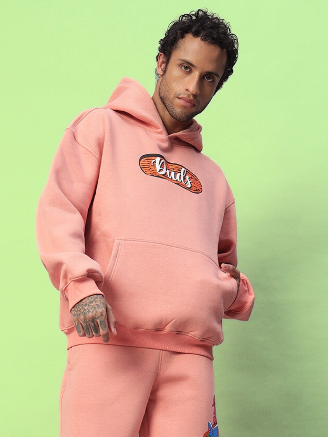 Man wearing a Deliberate Fleece Hoodie in peach color, featuring a front pocket and logo emblem, set against a green background.