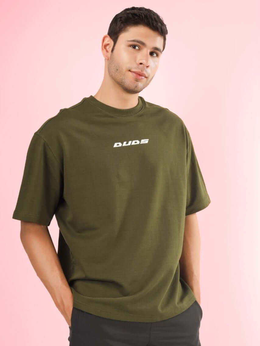 Man wearing AMOUR OVER-SIZED T-SHIRT OLIVE GREEN made with premium cotton and elastane, featuring a casual fit on a pink background.