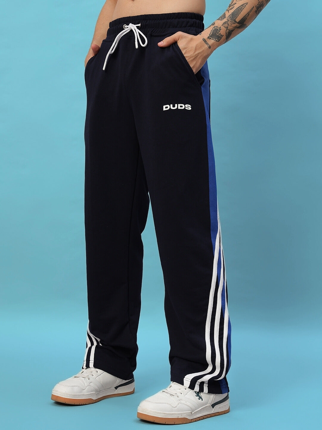 Man wearing navy blue TANGLE relaxed-fit joggers by DUDS with white sneakers in front of a blue background.