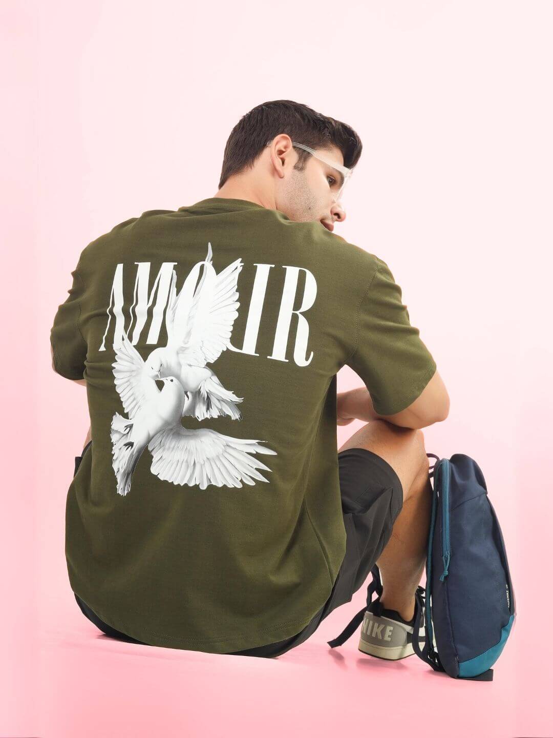 Man wearing AMOUR OVER-SIZED T-SHIRT OLIVE GREEN with white graphics on back, sitting against pink background.