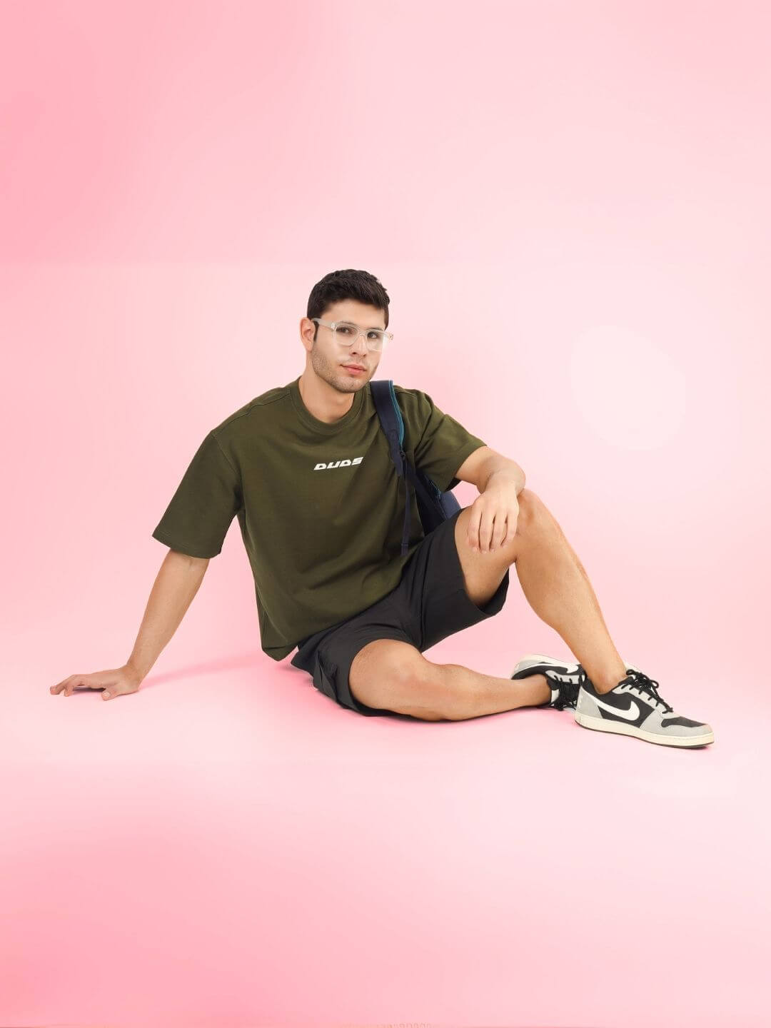 Model wearing AMOUR OVER-SIZED T-SHIRT OLIVE GREEN with premium heavy gauge fabric, sitting casually on pink background.