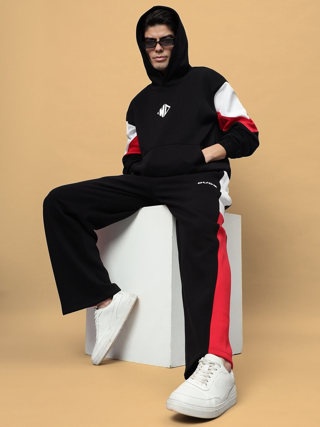 Person wearing BRIO Fleece Colorblock Co-Ord in black, featuring a stylish hoodie and joggers with red accents, sitting on a white block.