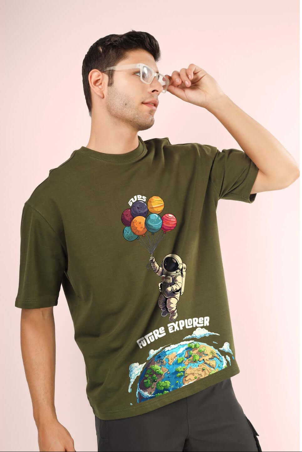 Man wearing FUTURE EXPLORER OVERSIZED T-SHIRT OLIVE GREEN with graphic print, standing casually with hand on glasses.