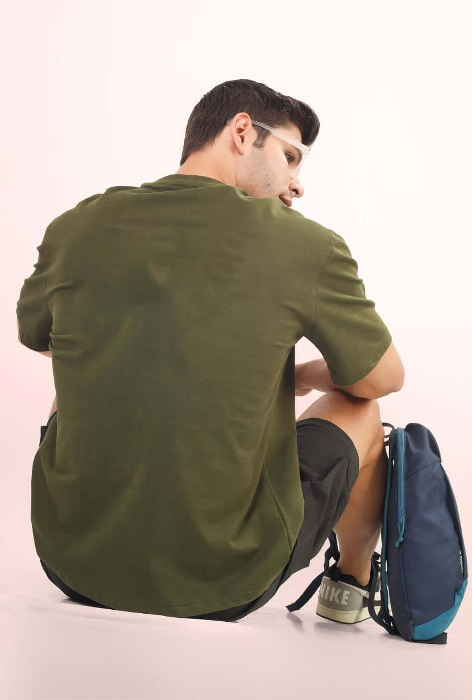 Back view of model wearing FUTURE EXPLORER OVERSIZED T-SHIRT OLIVE GREEN with a backpack on pink background.