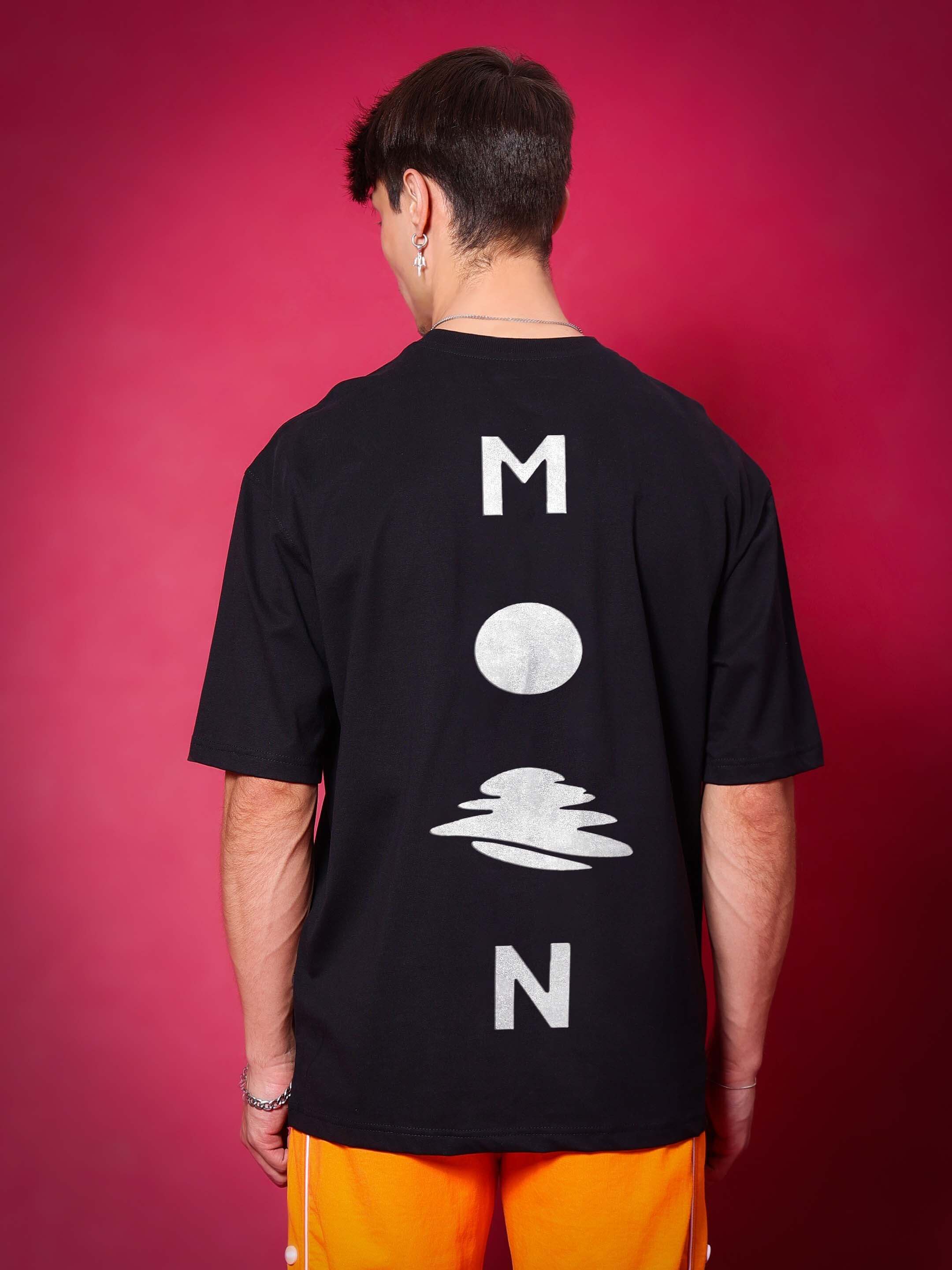 Person wearing black oversized t-shirt with glittered moon design on back, standing against pink background.