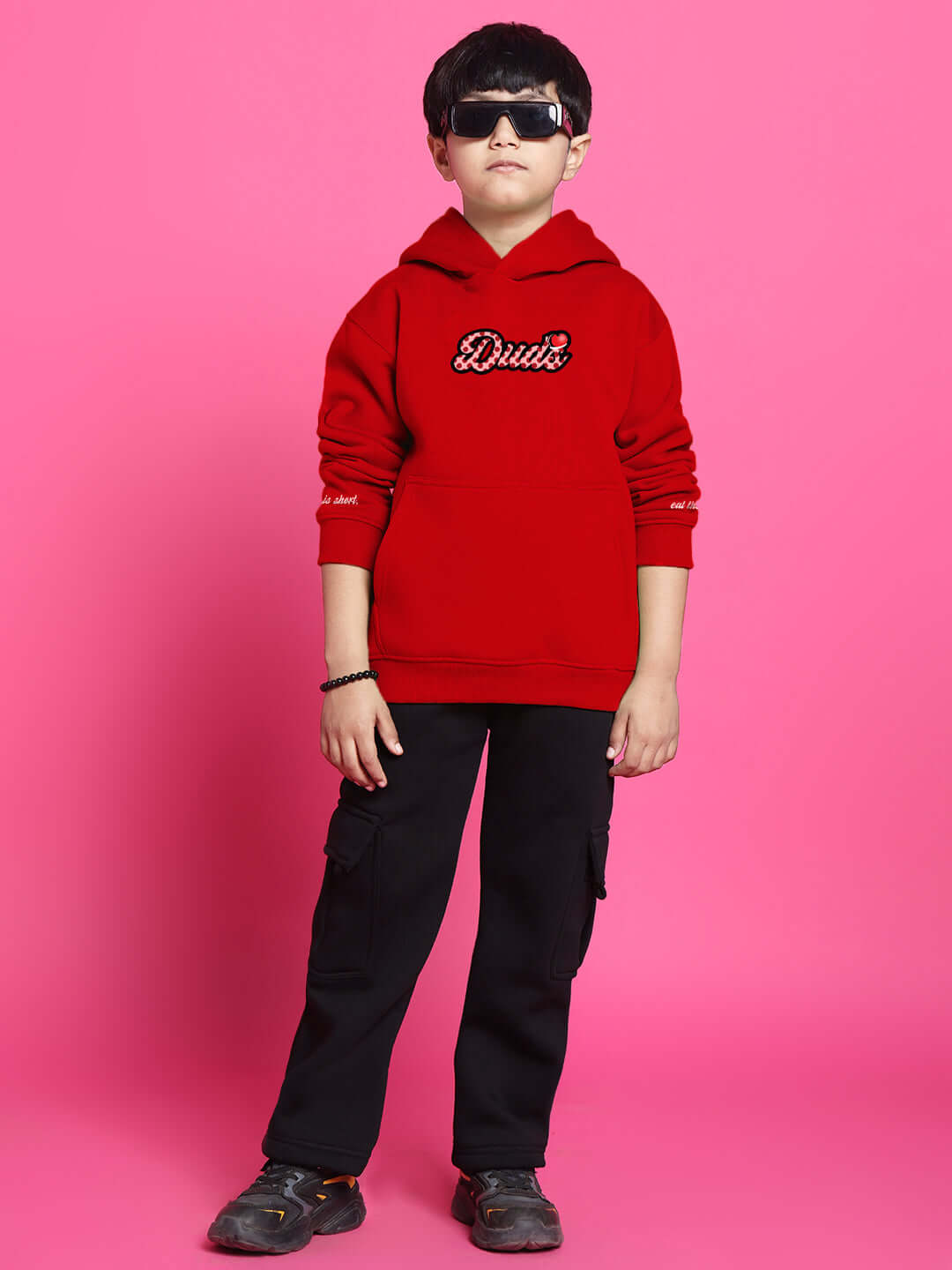 Child wearing a red hoodie and black pants co-ord set for boys and girls, standing against a pink background, showcasing casual style.