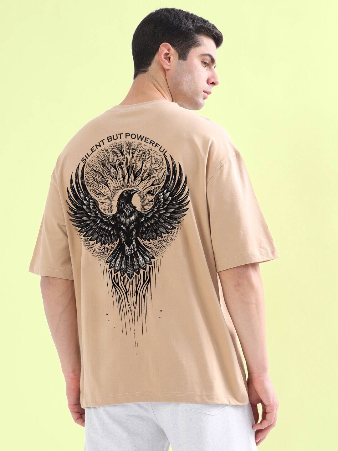 Man wearing SILENT BUT POWERFUL PRINTED T-SHIRT BEIGE with a bird design on back, standing against a yellow background