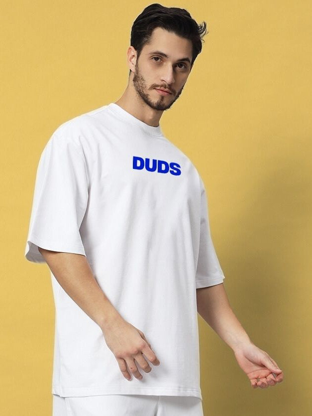 Man wearing ABSENT PRESENT OVER-SIZED T-SHIRT WHITE with blue logo, standing against a yellow background.