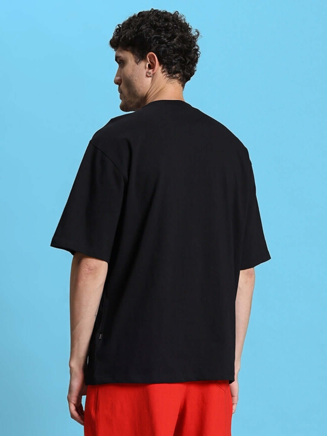Back view of man wearing ACRO oversized black T-shirt, premium heavy gauge 100% cotton, 200 GSM, styled with red pants against blue background.