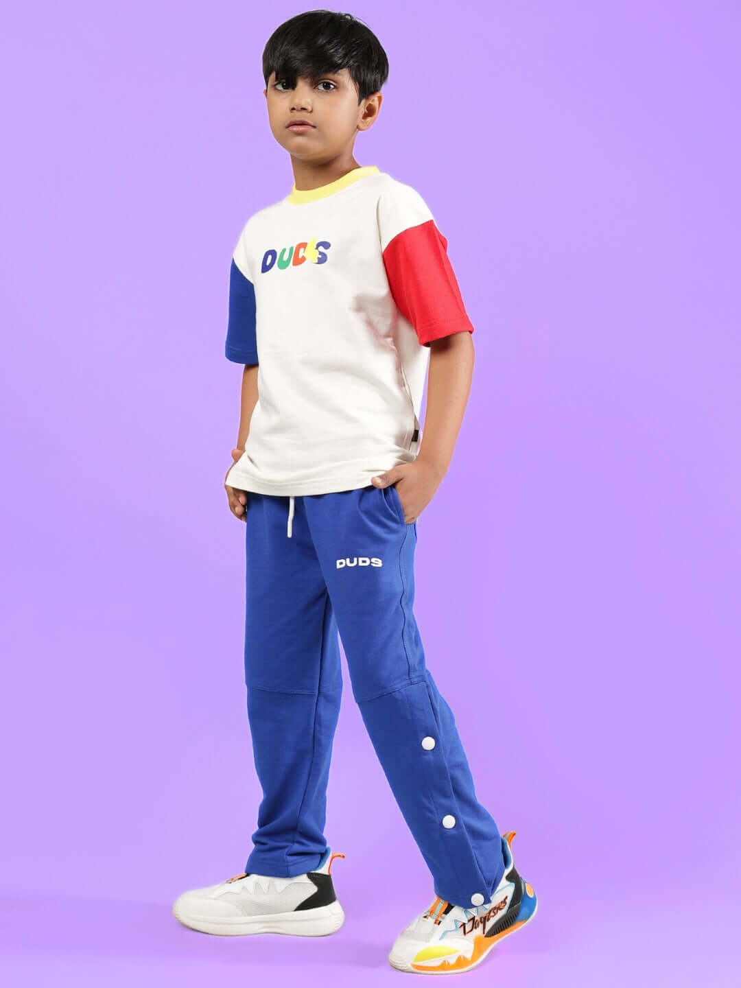 Child wearing Adonis Co-Ord outfit in off white and blue by DUDS, featuring color-blocked shirt and blue pants, against purple background.