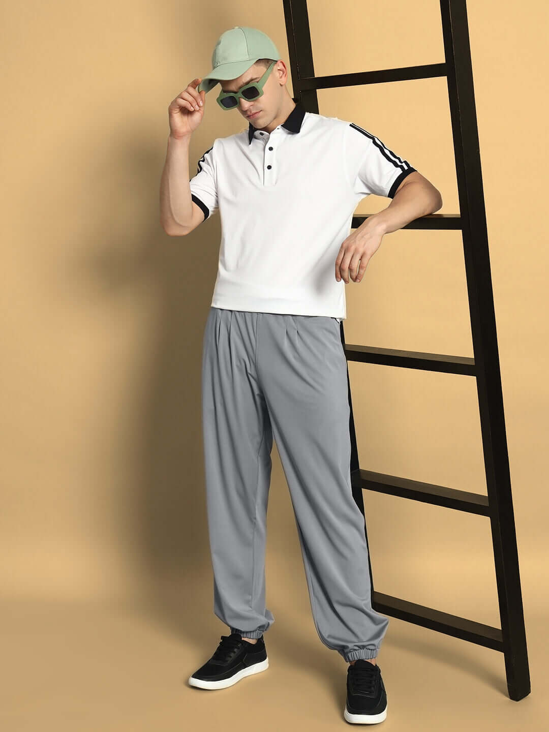 Man wearing AGILE RELAXED PANT JOGGER in grey, paired with a white polo shirt and cap, standing near a ladder in a stylish pose.