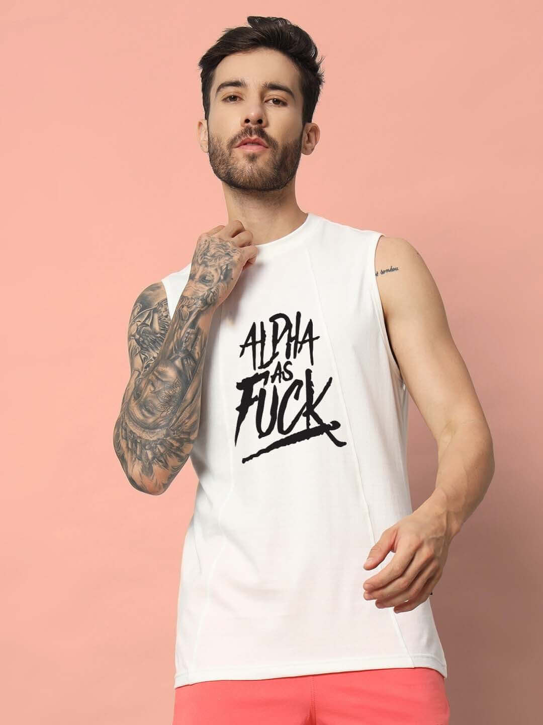 Man wearing off-white 'Alpha As Fuck' gym t-shirt, tattooed arm, pink shorts, against a pastel background.