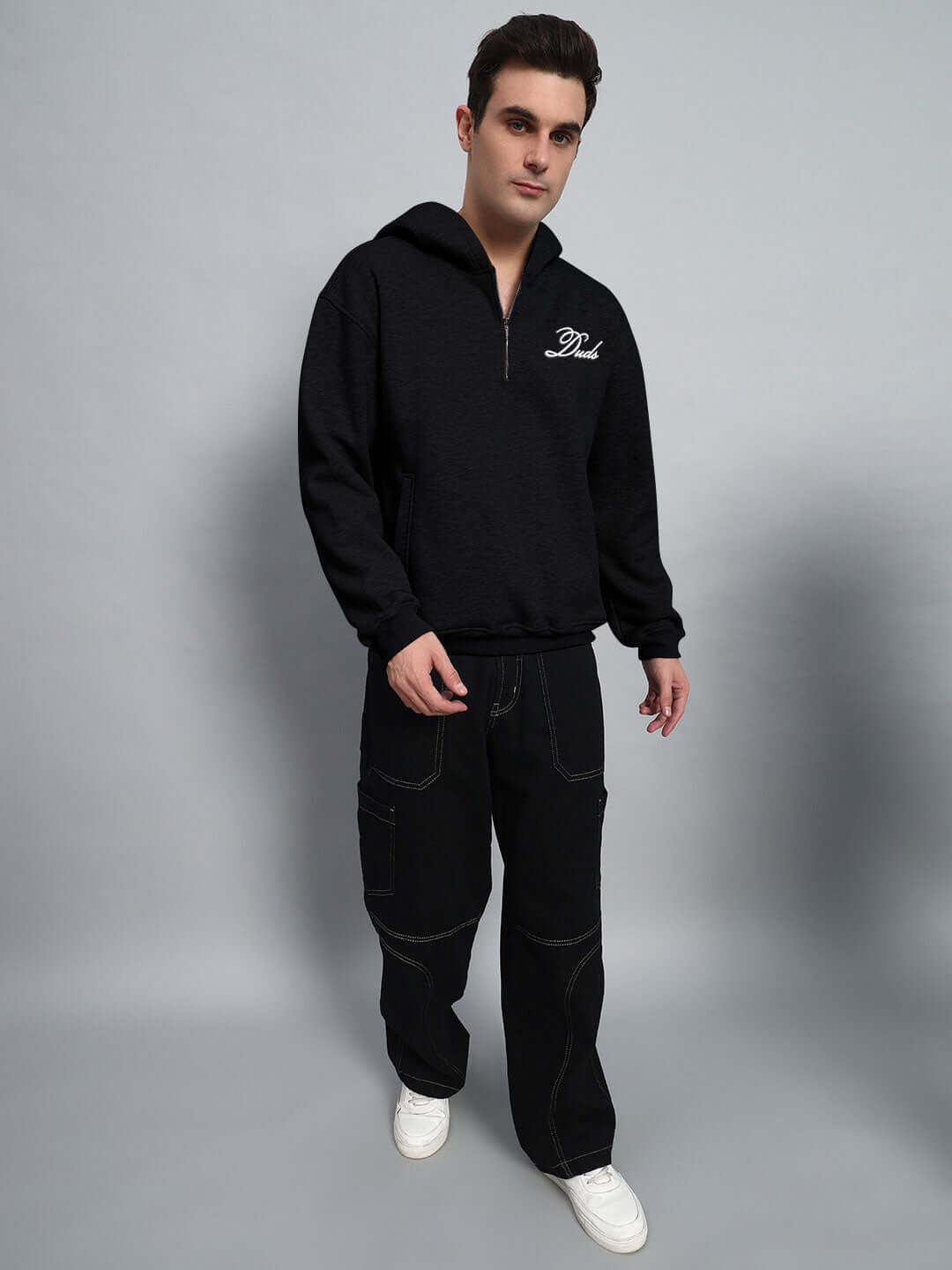 Man wearing WEARDUDS AMARILLO FLEECE HOODIE BLACK with white sneakers standing against gray background