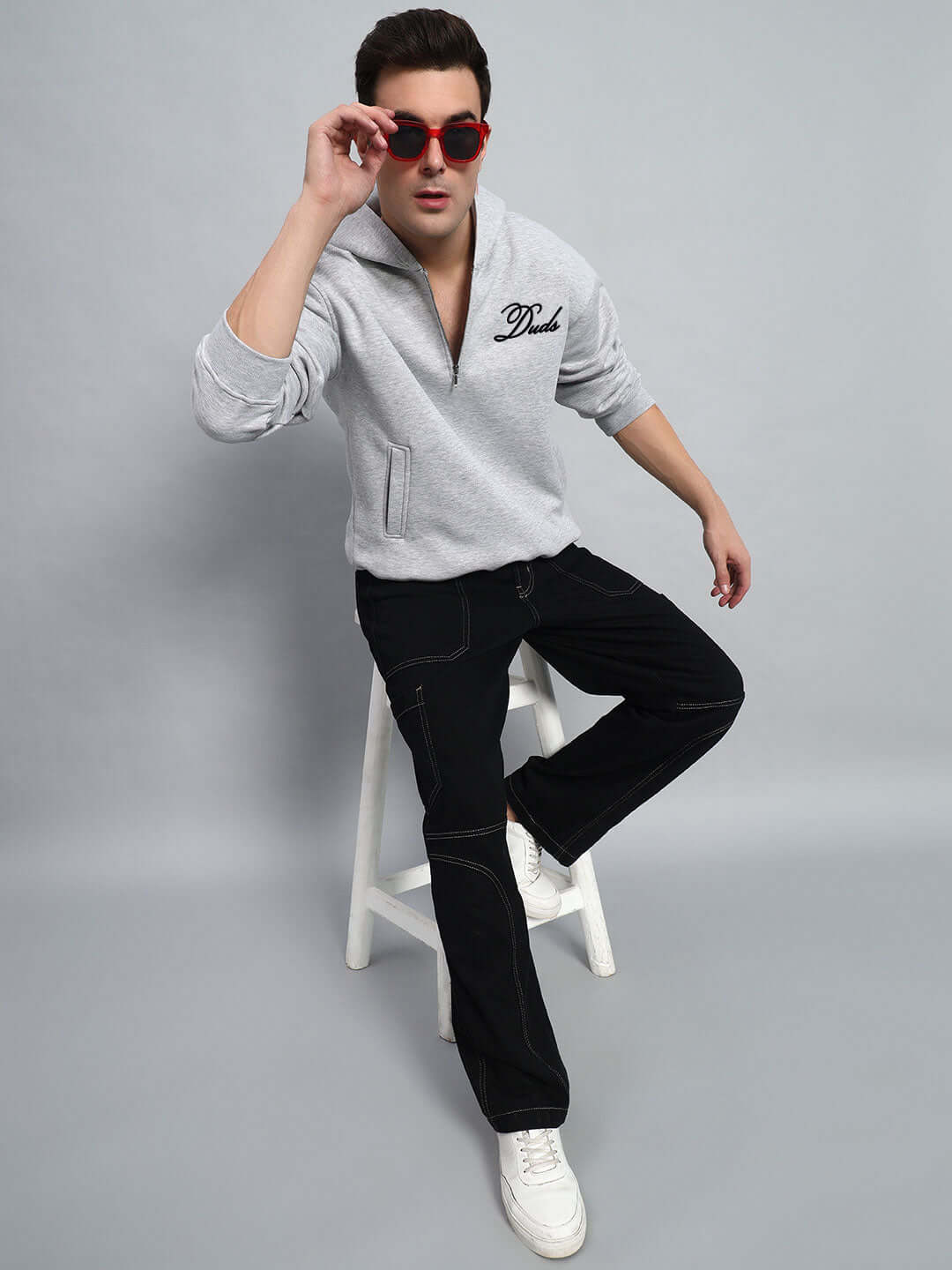 Model wearing AMARILLO FLEECE HOODIE MELANGE GREY with red sunglasses, seated on a stool, showcasing stylish design and fit.