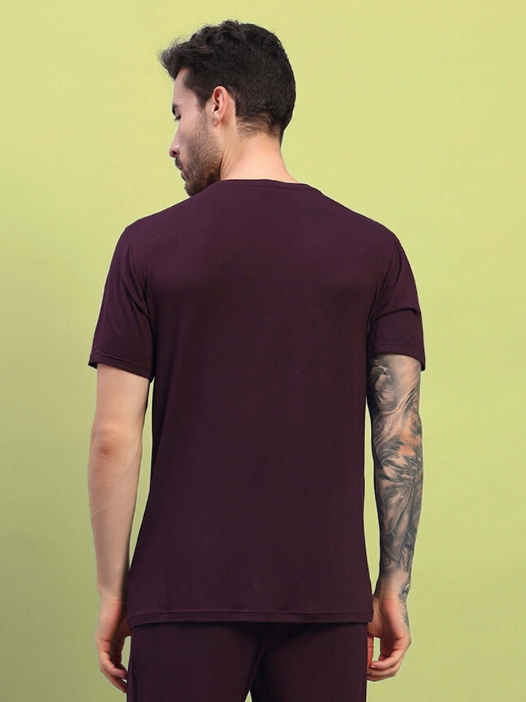 Man wearing maroon AMBLE T-shirt made of premium modal fabric, showcasing back view on a green background. Bio-washed and made in India.