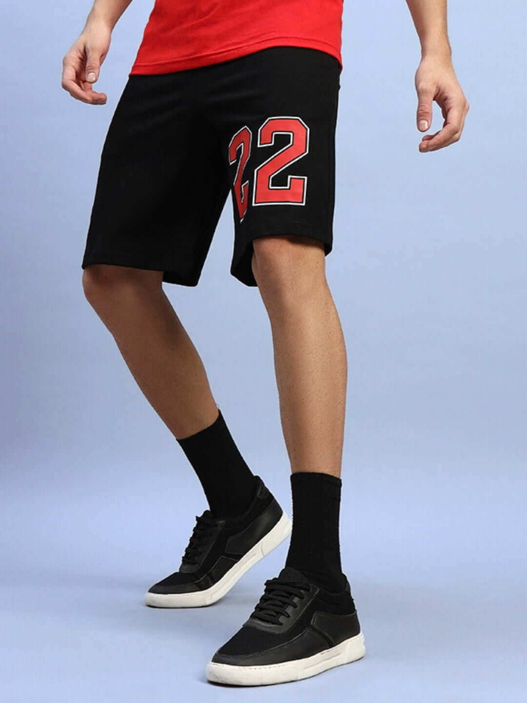 Man wearing AMIGA REGULAR FIT SHORTS BLACK with number 22 and black sneakers on a blue background.