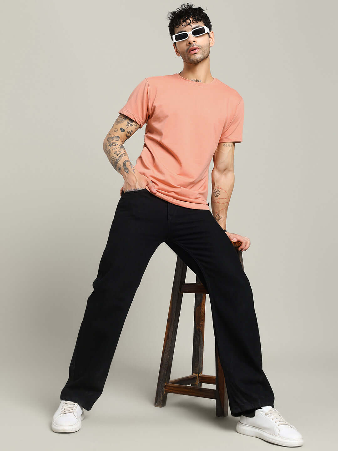 Model wearing Andrew Regular Fit T-shirt in peach, 100% cotton, stylish casual wear, sitting on stool, white sneakers, modern fashion.
